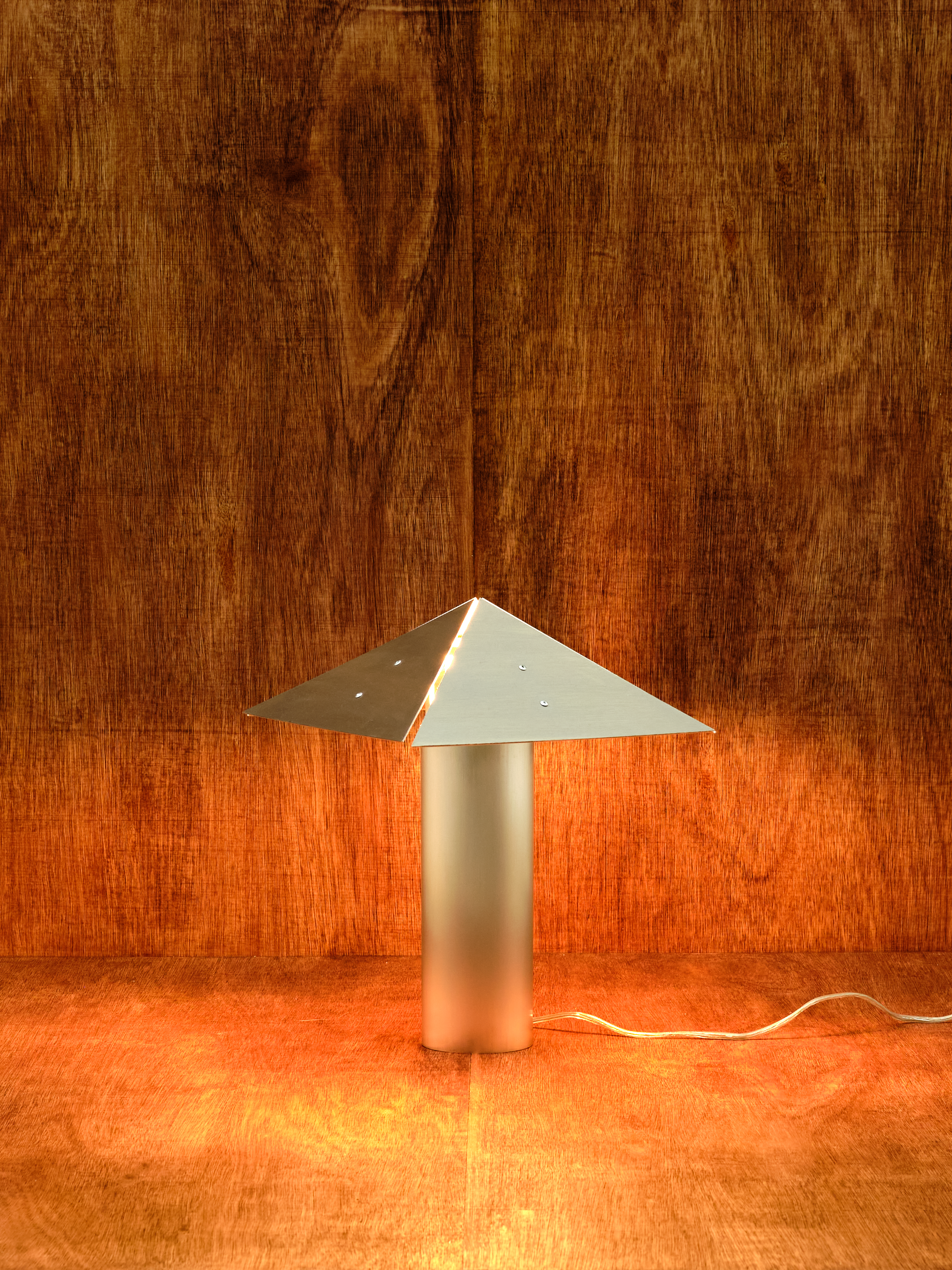 Metallic lamp with a pyramid-shaped design on a wooden floor and wall.